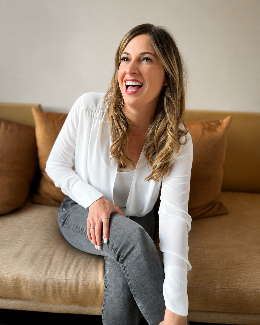 mary harcourt sitting on a brown couch laughing with a neutral background