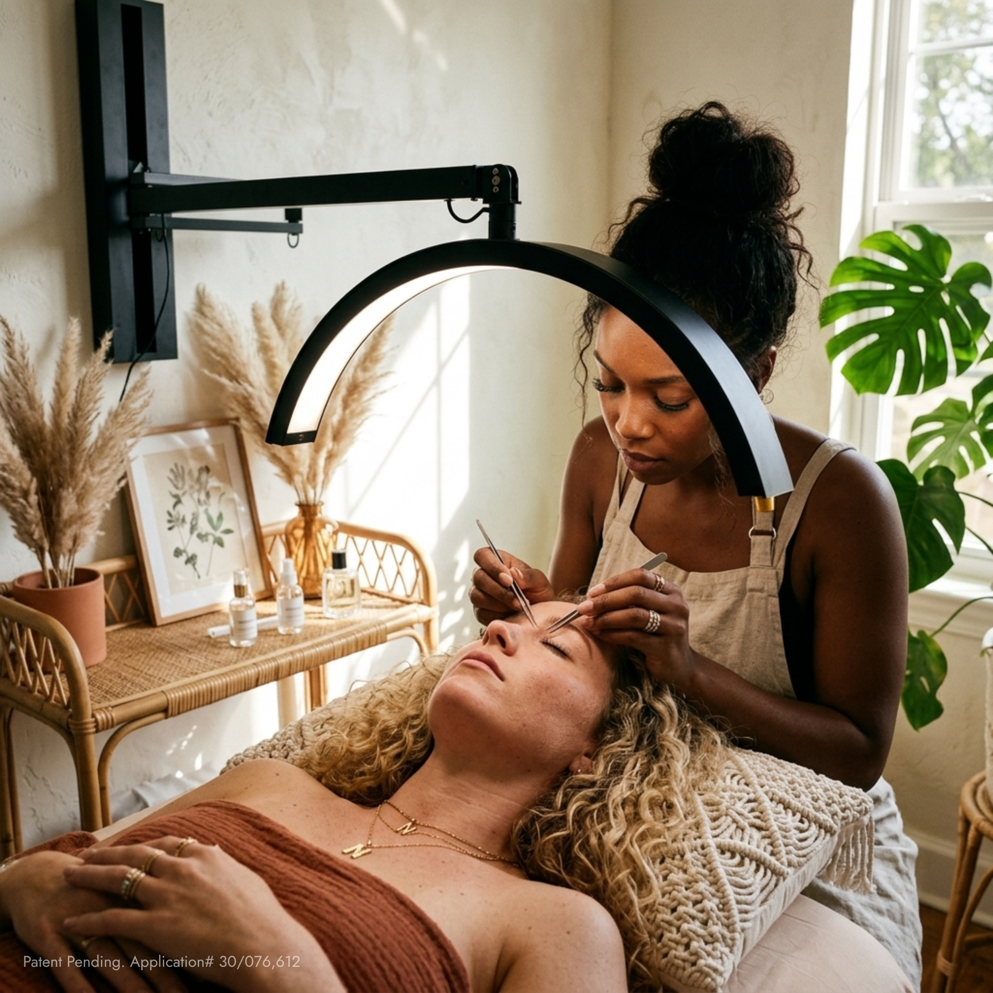 A black LED eyebrow microblade treatment lamp mounted on a wall, illuminating a woman receiving eyebrow tattooing while another woman attends to her. The scene is set in a bright, modern spa room with natural light, a rattan table with dried pampas grass, and a green plant in the background.