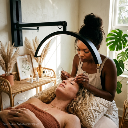 A black LED eyebrow microblade treatment lamp mounted on a wall, illuminating a woman receiving eyebrow tattooing while another woman attends to her. The scene is set in a bright, modern spa room with natural light, a rattan table with dried pampas grass, and a green plant in the background.