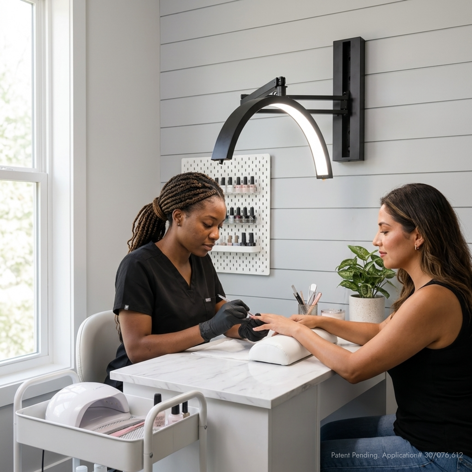 A nail salon scene with a manicurist wearing black scrubs and gloves working at a white manicure table under a black wall-mounted arc lamp, while a client sits at the table. The room has light gray shiplap walls, a window on the left, and a small plant on the counter.