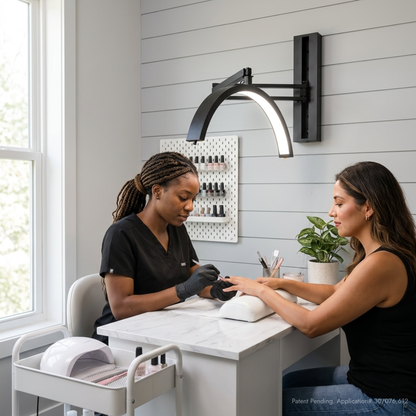 A nail salon scene with a manicurist wearing black scrubs and gloves working at a white manicure table under a black wall-mounted arc lamp, while a client sits at the table. The room has light gray shiplap walls, a window on the left, and a small plant on the counter.