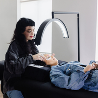 Woman applying eyelashes to another woman using a lamp in a clinical setting.