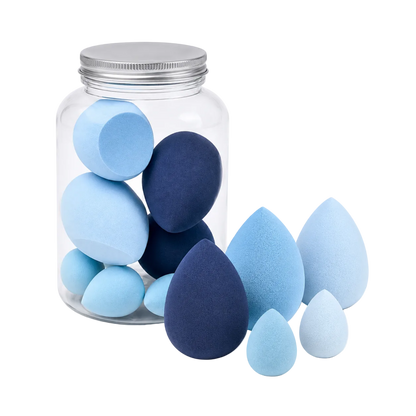 A set of makeup sponges in light blue, navy blue, and white, with some arranged inside a clear glass jar with a silver lid and others standing outside on a white background.