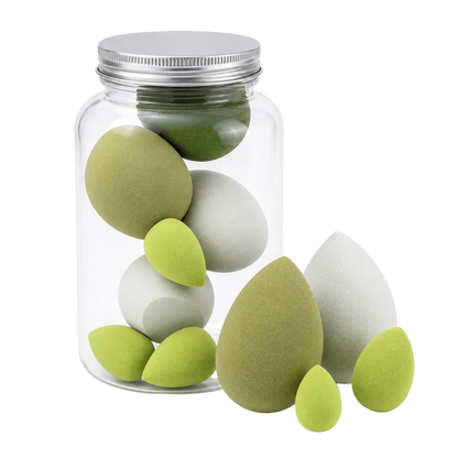 A clear glass jar with a silver metal lid containing several teardrop-shaped makeup sponges in light green and white, with three additional sponges placed in front on a clean white background.