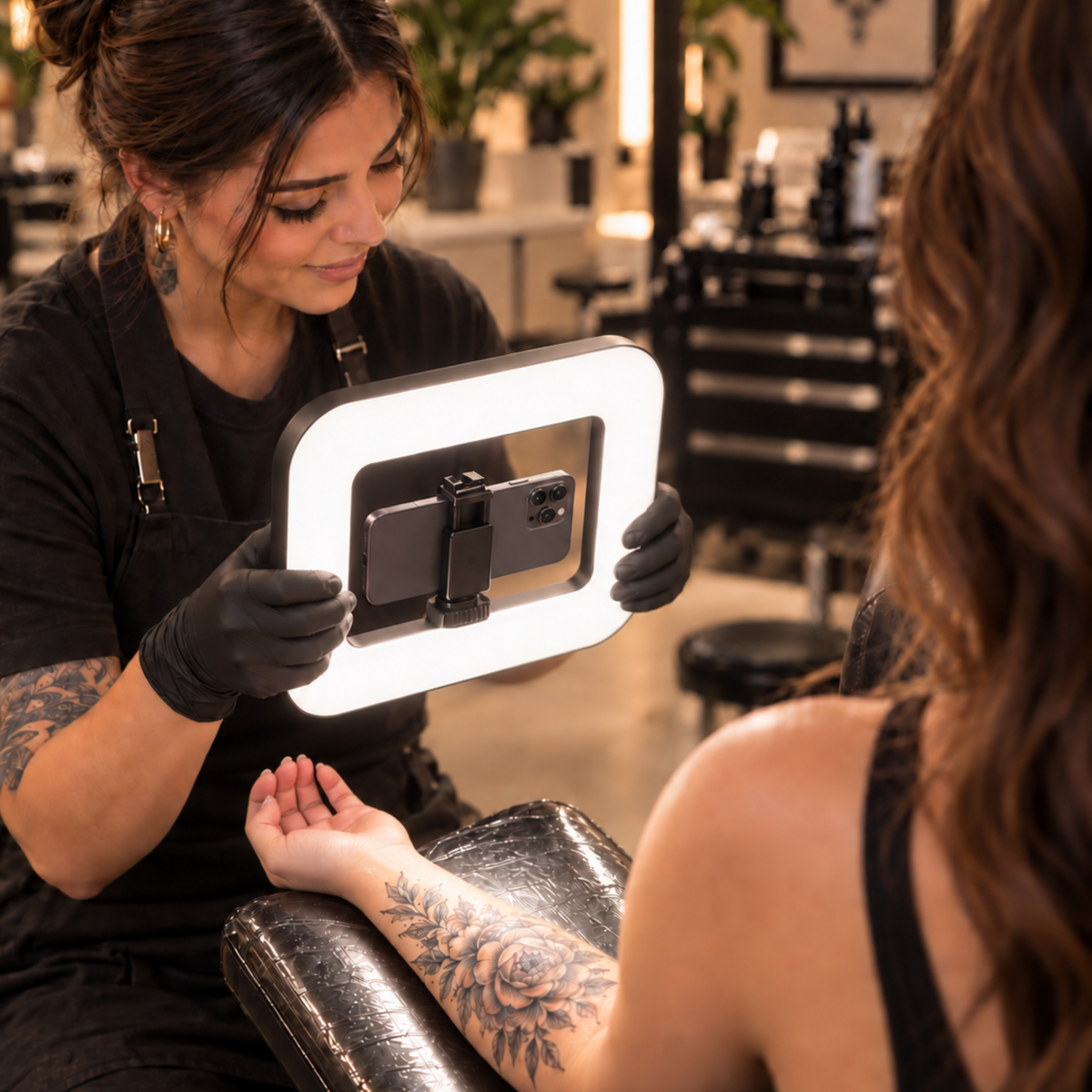 A tattoo artist wearing black gloves holds a rectangular ring light with a phone mounted inside, illuminating the artist&