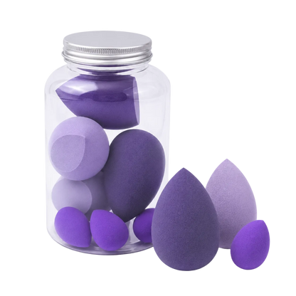 A clear glass jar with a silver metal lid containing multiple teardrop-shaped makeup sponges in shades of purple and lavender, with three additional sponges placed outside the jar on a white background.