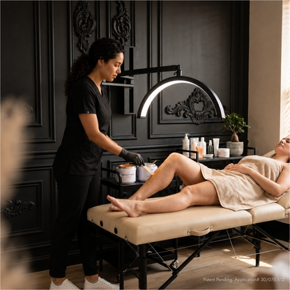 A spa treatment scene with a therapist in black scrubs attending to a client lying on a beige massage table. The room has dark decorative wall paneling, a curved ring light, and skincare products on a table.