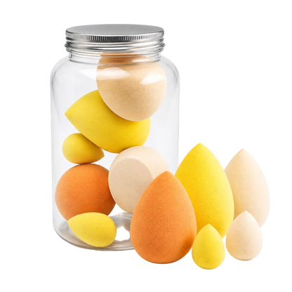 A clear glass jar with a silver metal lid containing multiple teardrop-shaped makeup sponges in yellow, orange, and beige, with several sponges placed in front on a white background.